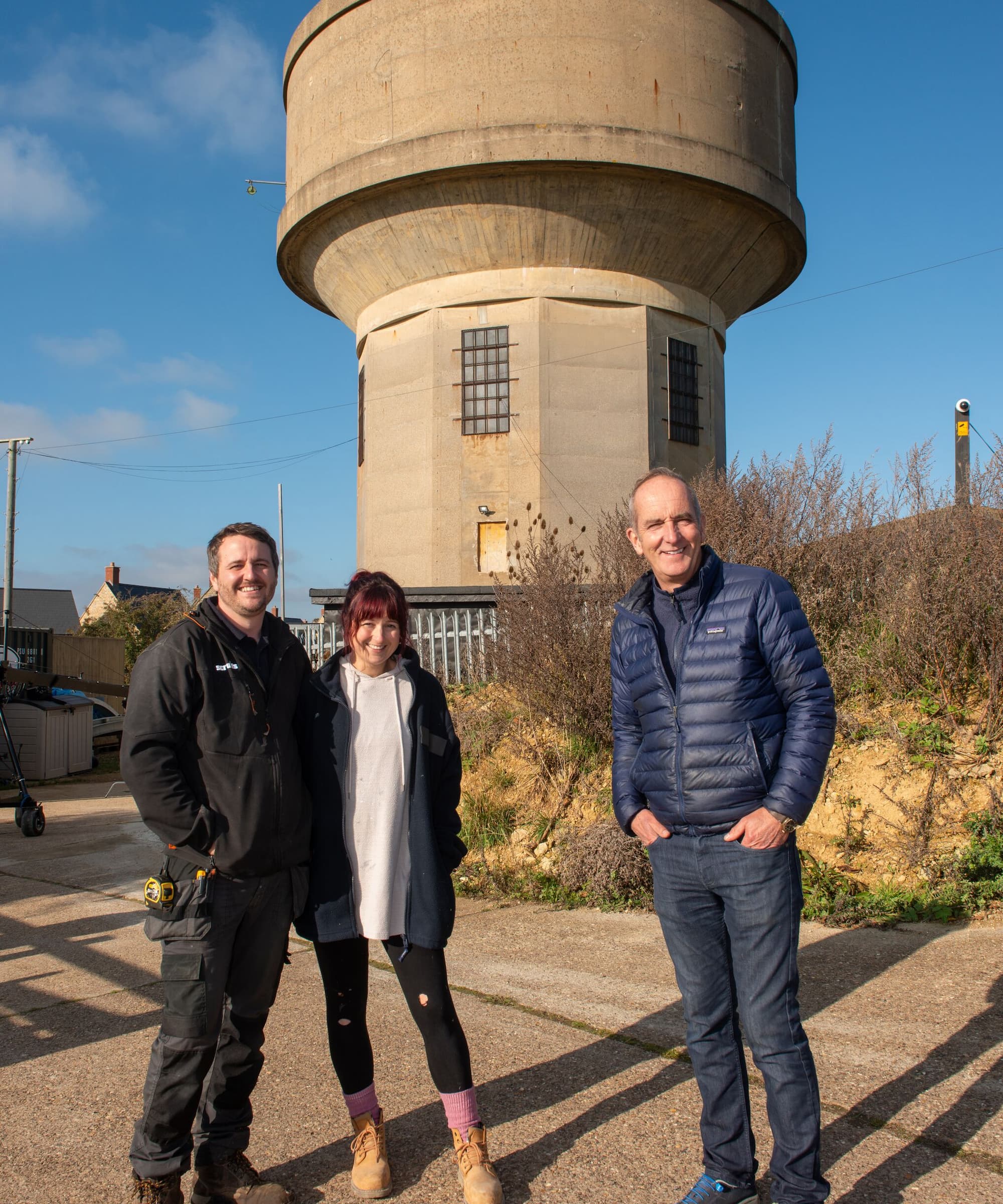 Kevin McCloud and a couple stood in front a massive water tower with a metal fence in front of it