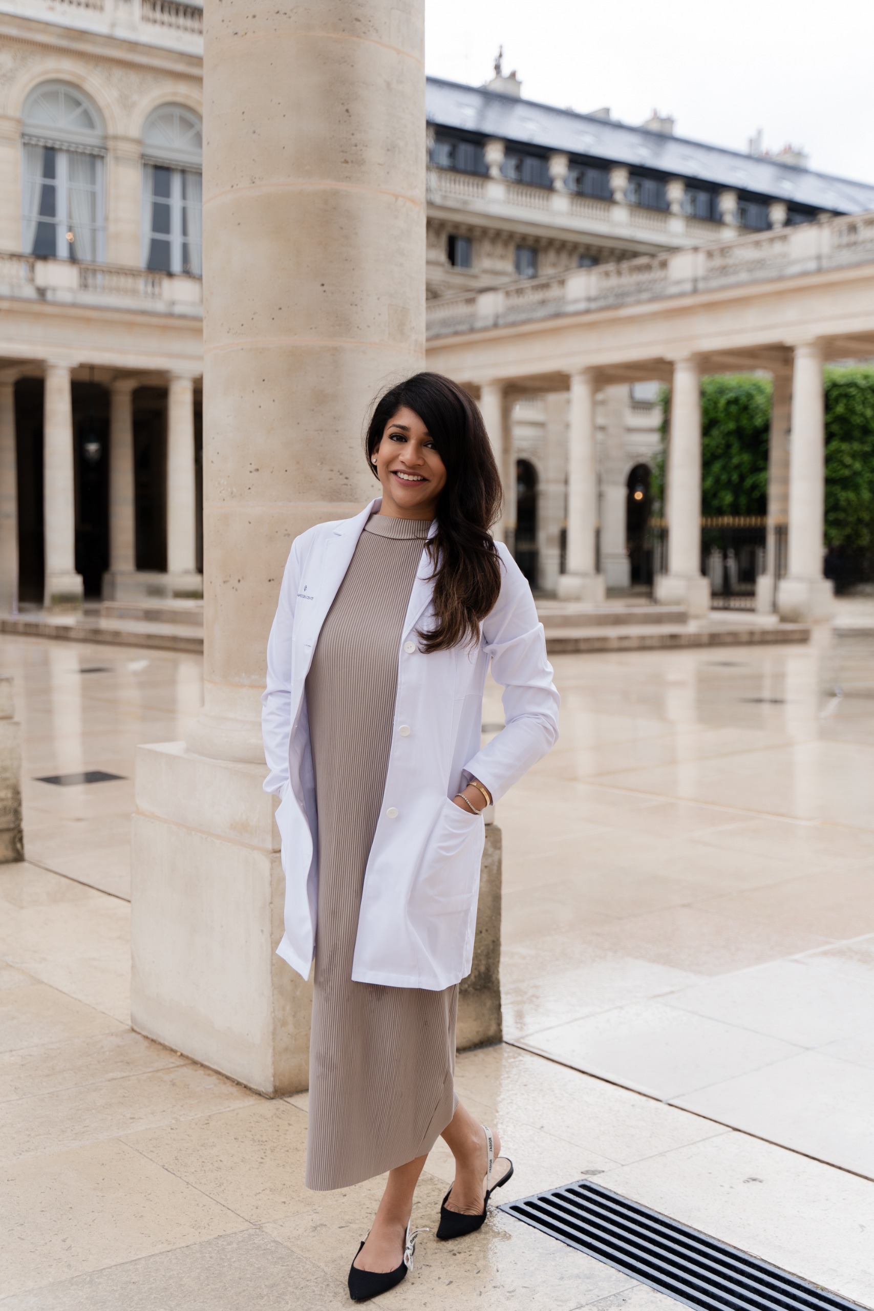 a woman in a tan dress and white lab coat with black hair and black heels