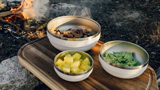 Three Yeti Rambler bowls with food in them and a campfire in the background