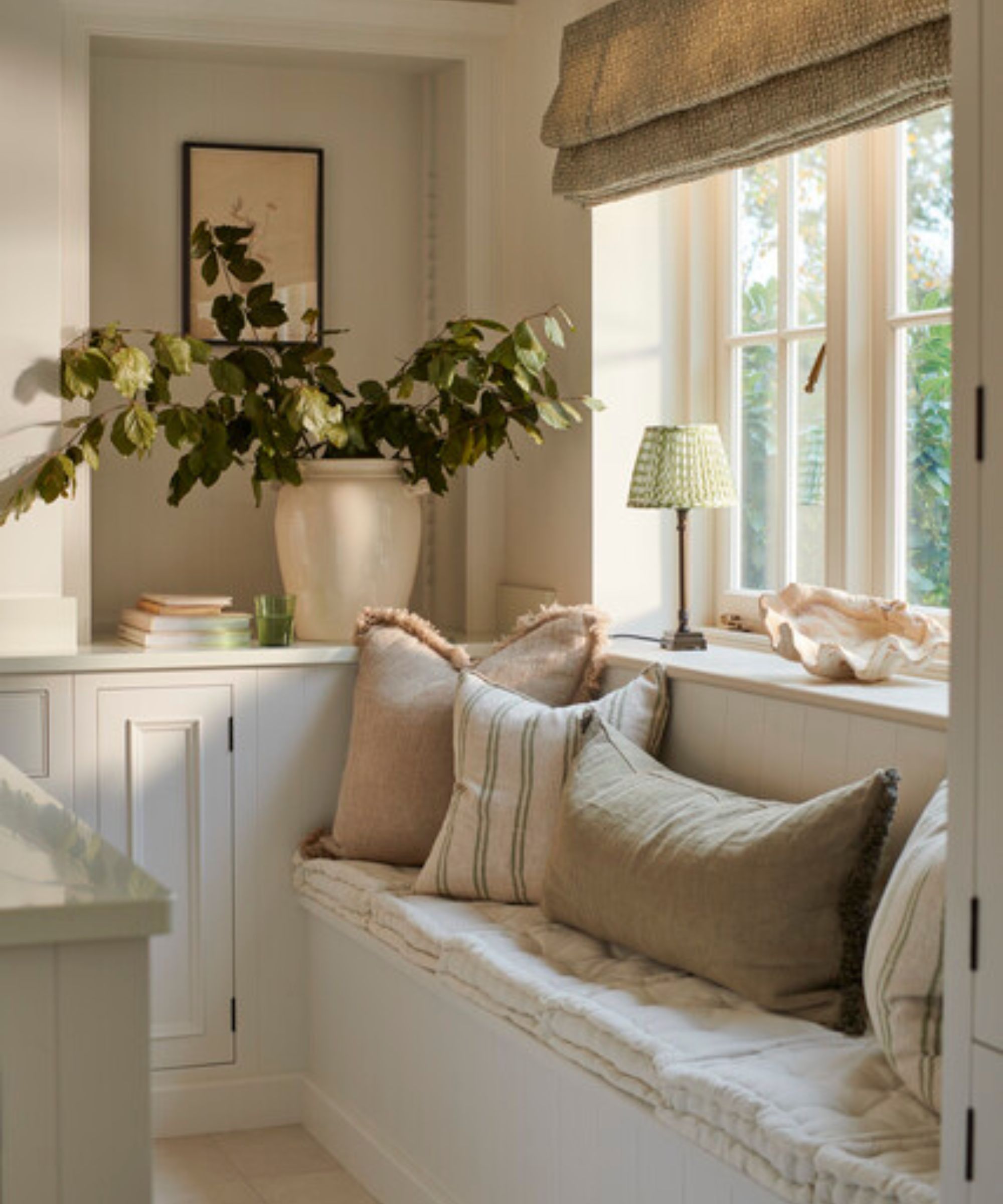 A sociable kitchen with a cozy window seat dressed with throw pillows, a vase of branches, and a table lamp