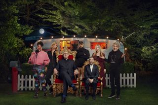 A group shot of the series 12 Taskmaster contestants outside the Taskmaster caravan: Desiree Burch, Morgana Robinson, Guz Khan, Victoria Coren Mitchell and Alan Davies. Greg Davies and Alex Horne are sitting in front in their thrones, Alex has been digitally manipulated to appear obviously smaller than Greg.