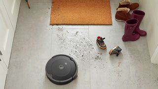 iRobot Roomba 692 vacuum cleaner shown on tile floors near a doormat, with mud on the floor and two pairs of shoes.