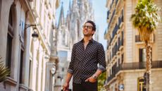 A well-dressed middle-aged man walks through the old section of a European city with his roller suitcase. He is clearly happy.