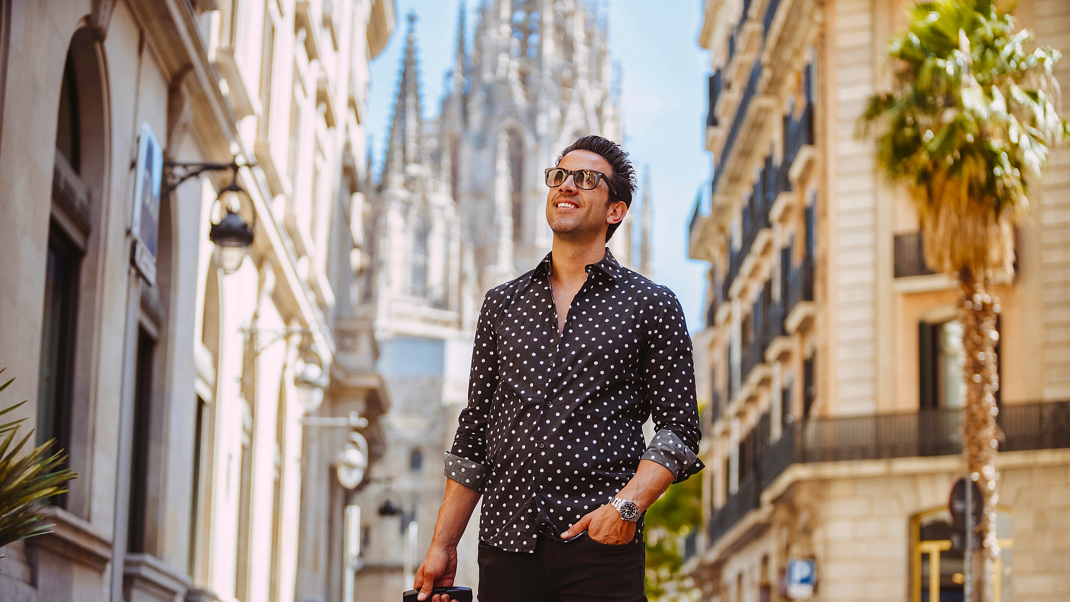 A well-dressed middle-aged man walks through the old section of a European city with his roller suitcase. He is clearly happy.