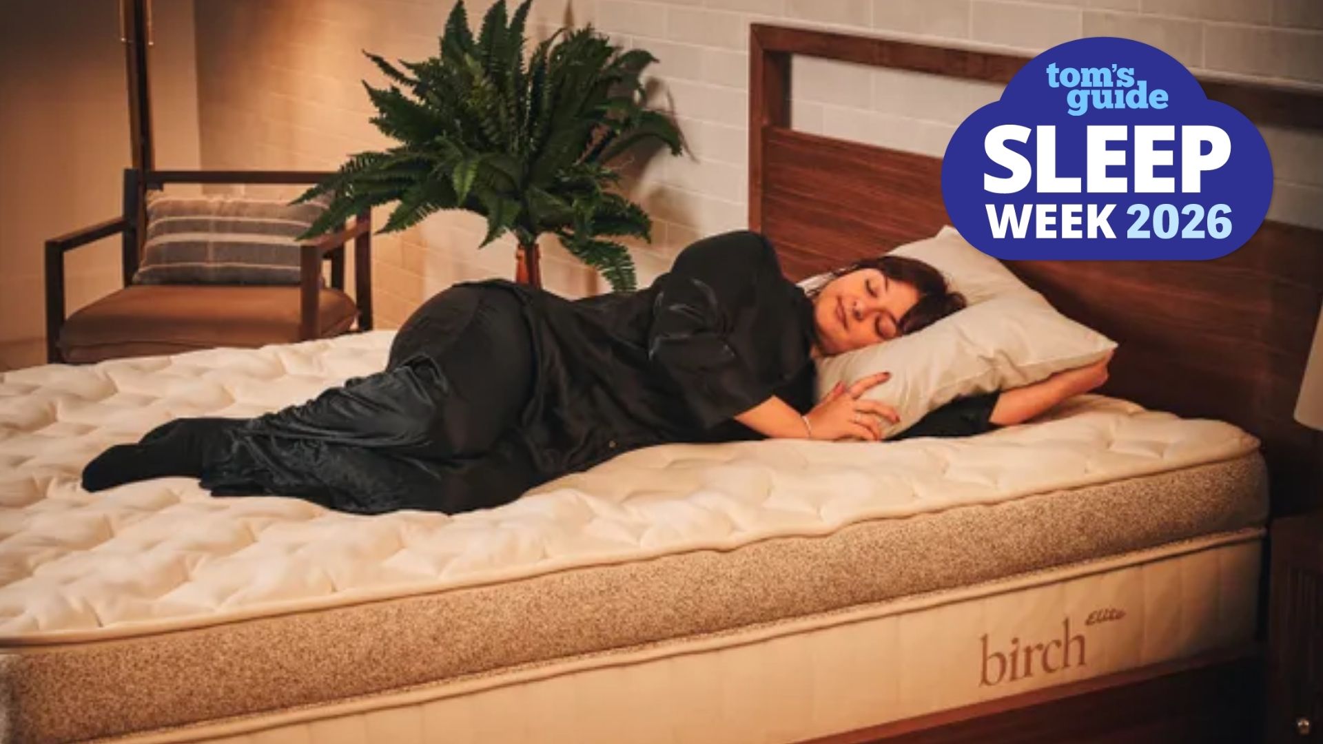 The image shows a dark haired woman lying on her side on a beige and brown mattress, placed on a wooden bed frame in a neutrally decorated bedroom. A blue cloud-shaped Tom's Guide Sleep Week 2026 is overlaid on top of the image in the bottom right hand corner.