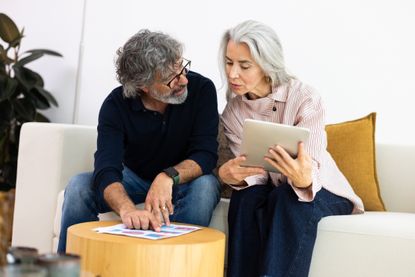 Mature couple sitting on sofa, planning budget and investments with tablet and financial documents