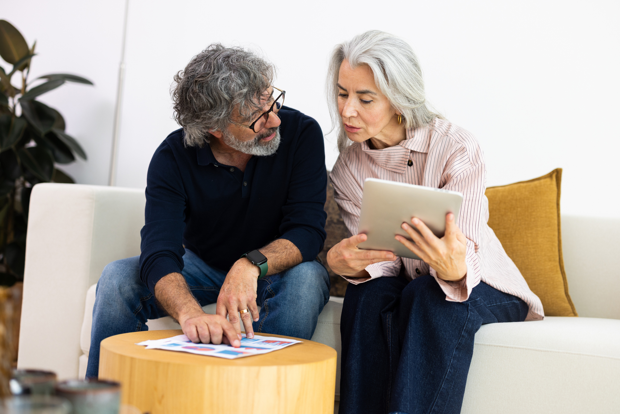 Mature couple sitting on sofa, planning budget and investments with tablet and financial documents