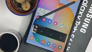 En Samsung Galaxy Tab S6 Lite ligger på sin förpackning bredvid en liten kruka och en kaffekopp, med en stylus liggandes på skärmen.