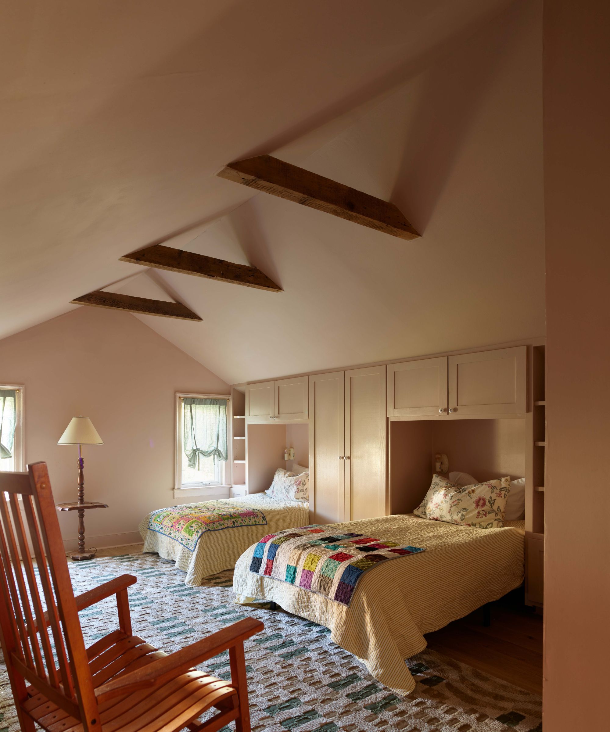 Attic bedroom with a sloped ceiling, exposed wooden beams, two twin beds recessed into built-in cabinets, and a wooden rocking chair in the foreground