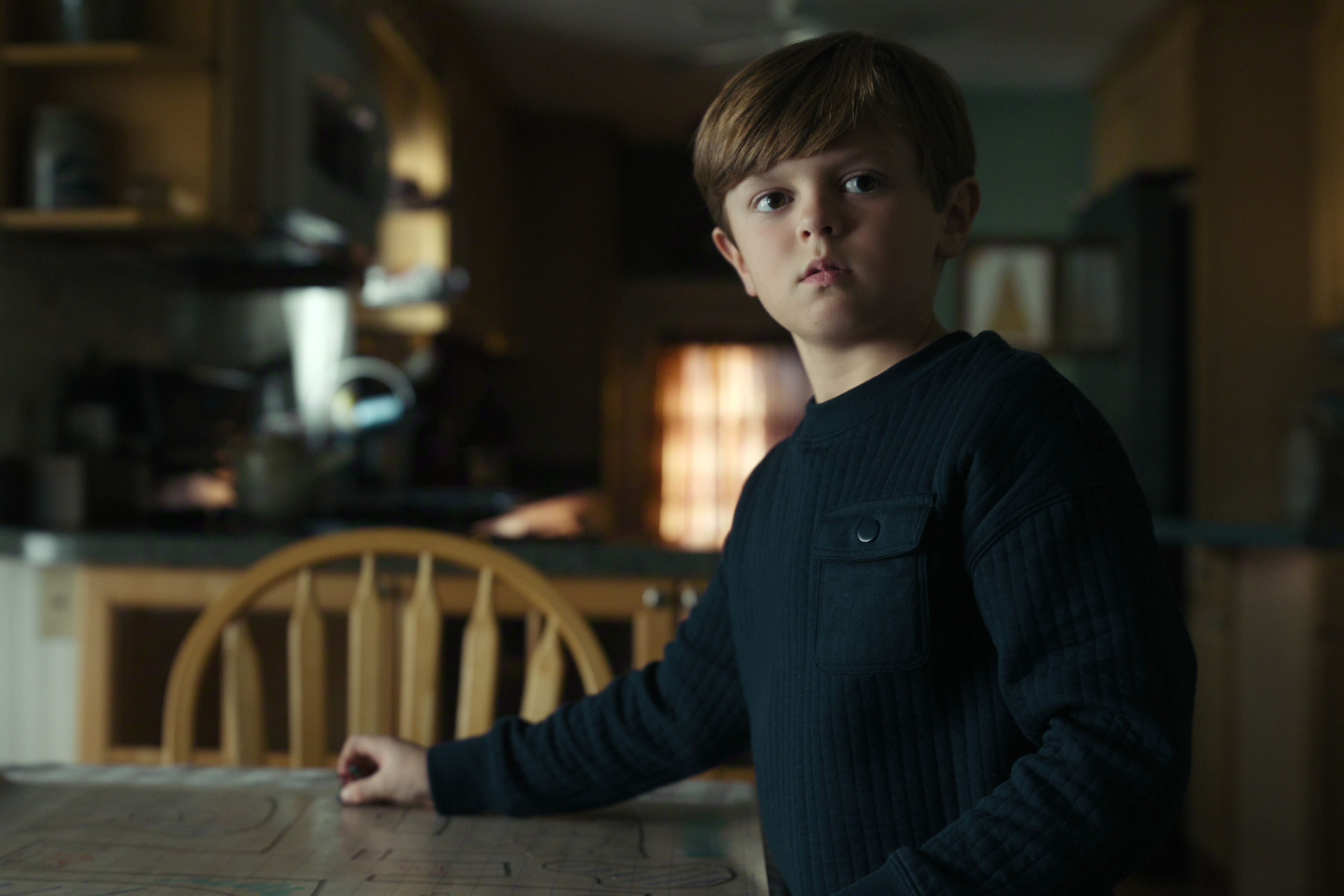 Child actor callum Vinson as The Son a small boy wearing a black sweater standing at a kitchen table in a still from the night agent season 3
