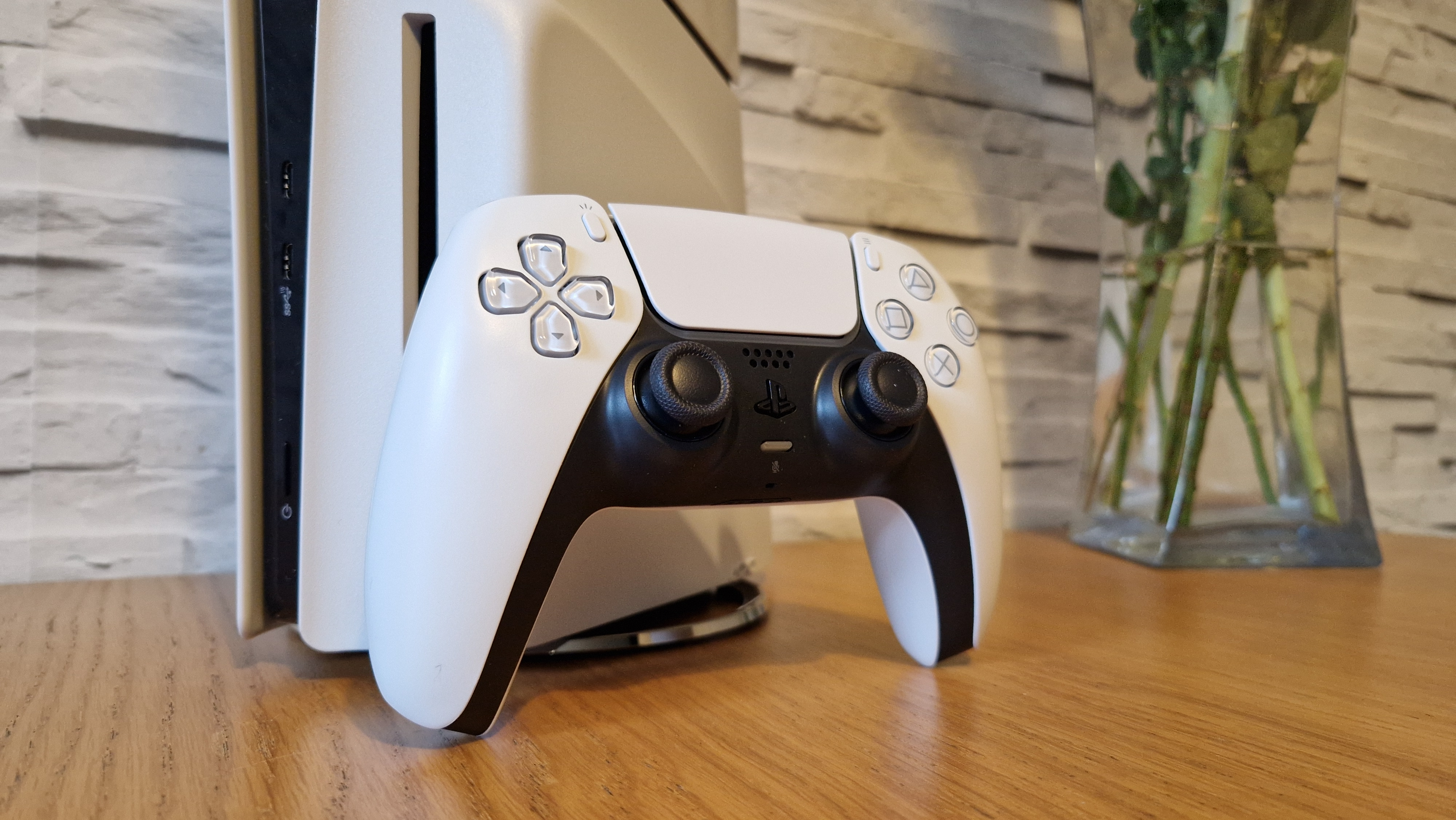 The PS5 Slim standing upright with a DualSense controller sitting on wooden table next to a vase of flowers with a white brick background