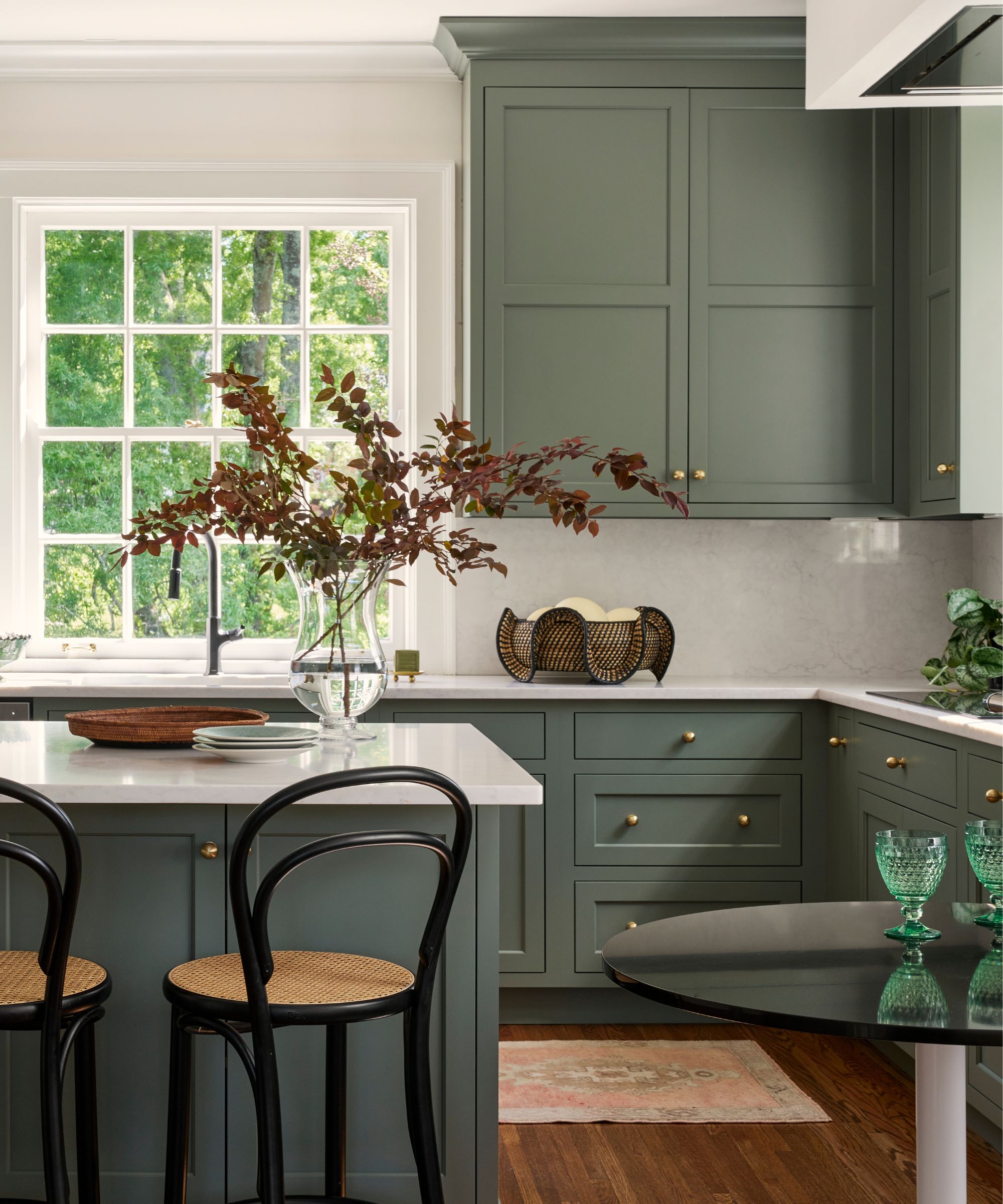 A modern kitchen with dark gray-green cabinets and a kitchen island, white walls and countertops, wood flooring, and a large window by the sink.