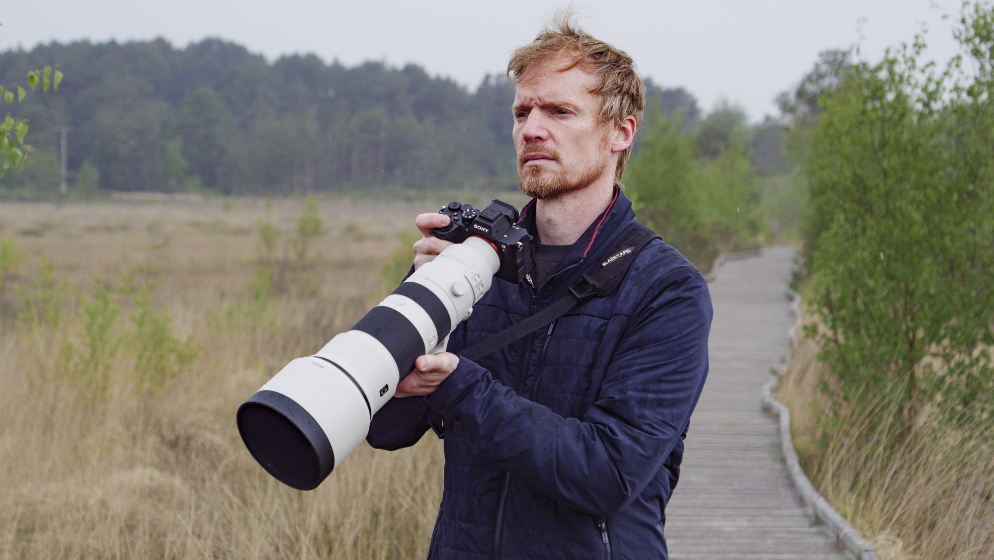 Man holding the Sony FE 400-800mm F6.3-8 G OSS supertelephoto zoom lens, he's standing on a boardwalk in a nature reserve