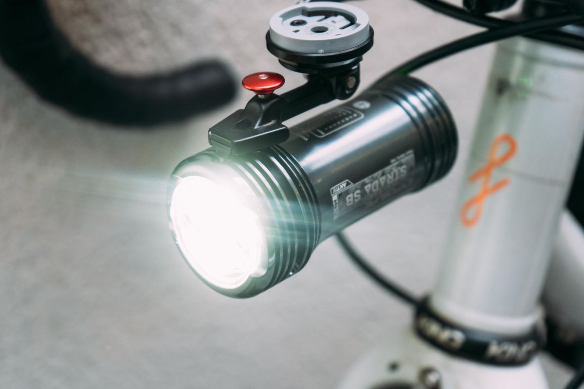 A silver bike light mounted underneath a bike computer mount and switched on