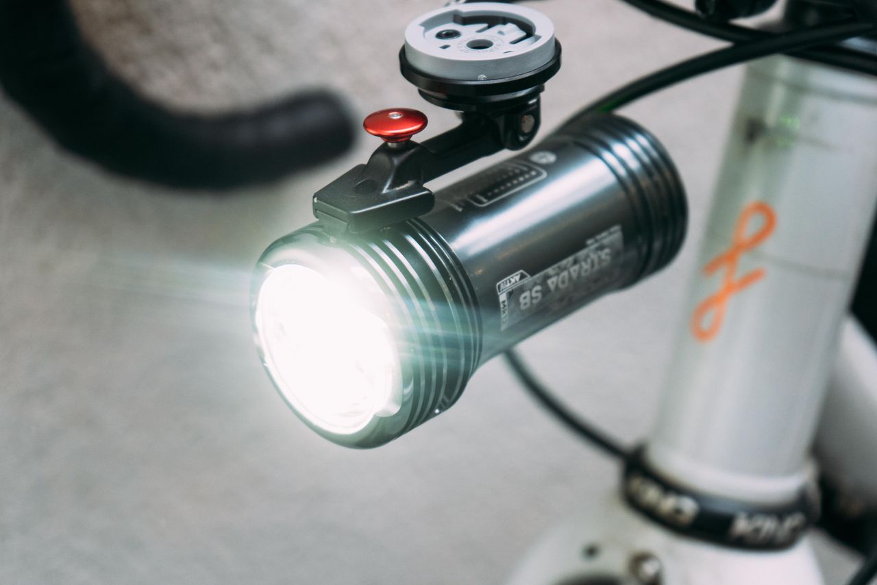 A silver bike light mounted underneath a bike computer mount and switched on