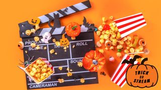 Film slate covered in popcorn, pumpkins, eyeball gummies and ghost ornaments on an orange background for Halloween