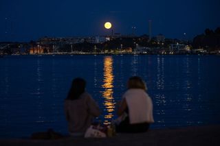 two people sit and look at a full moon shining over a large body of water with buildings and trees in the distance.