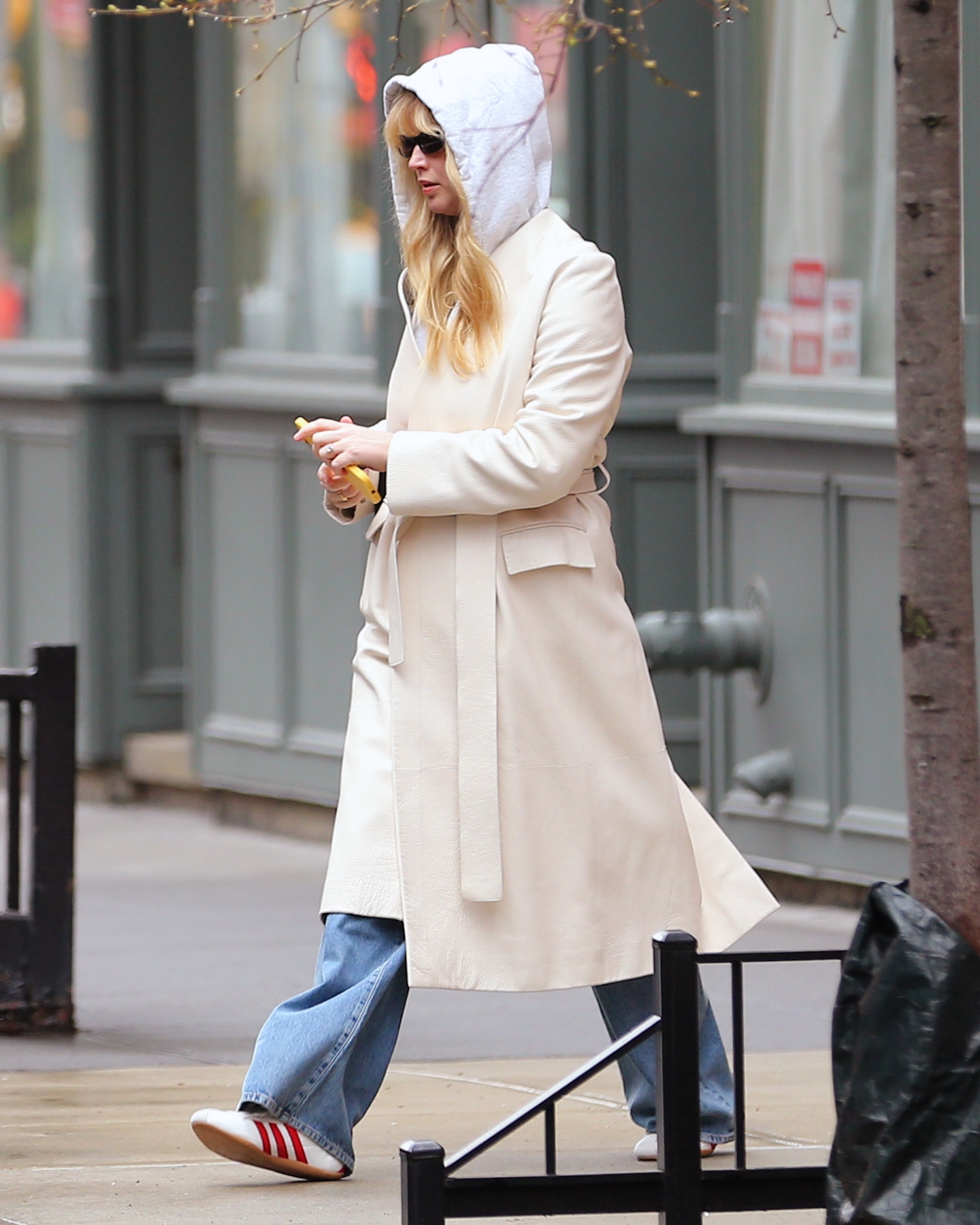Jennifer Lawrence walking in New York City wearing a gray hooded sweatshirt, cream belted trench coat relaxed, and Adidas Tokyo red-stripe sneakers.