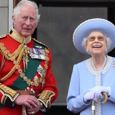 King Charles wears a red military uniform and looks concerned while standing on Buckingham Palace balcony with Queen Elizabeth II, who wears a baby blue outfit