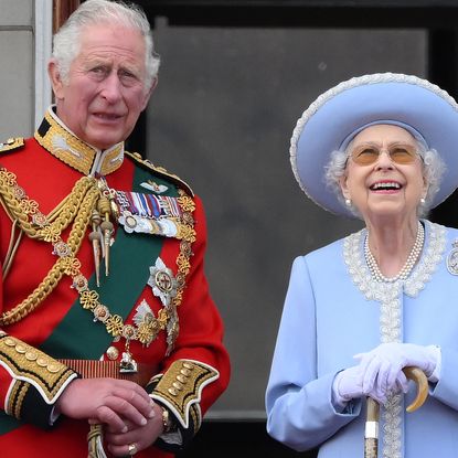 King Charles wears a red military uniform and looks concerned while standing on Buckingham Palace balcony with Queen Elizabeth II, who wears a baby blue outfit