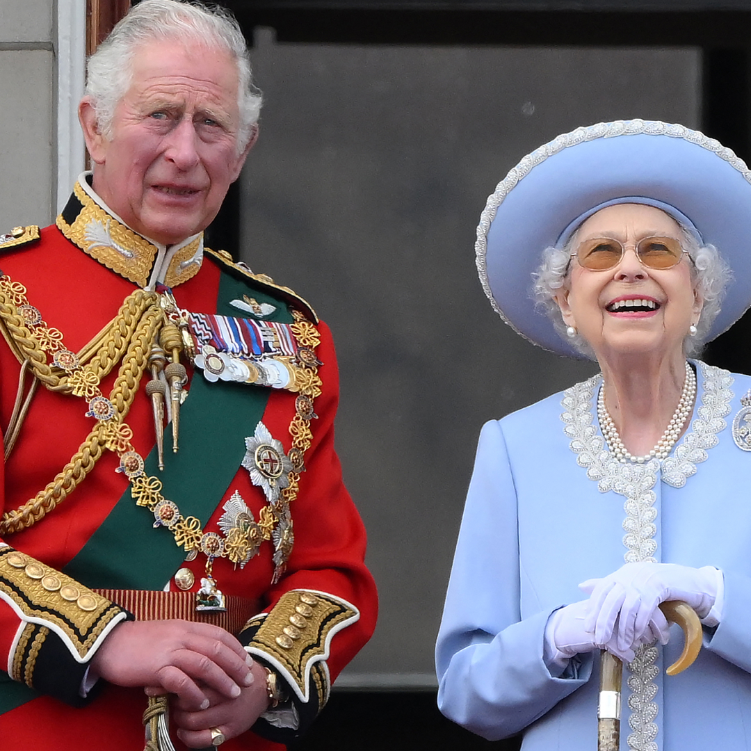 King Charles wears a red military uniform and looks concerned while standing on Buckingham Palace balcony with Queen Elizabeth II, who wears a baby blue outfit