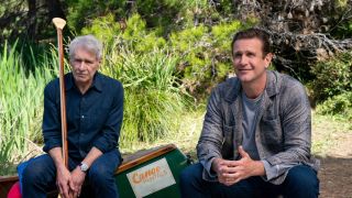 From left to right: Harrison Ford looking glum while holding an ore and Jason Segel sitting next to him, looking up and smiling. 