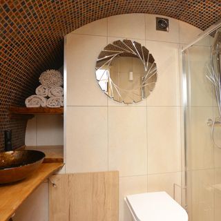 hobbit house bathroom with earthy mozaic tiles along the curved wall and ceiling, large white tiles along the back wall, a circle feature mirror, chrome sink on wooden unit along with a toilet and shower