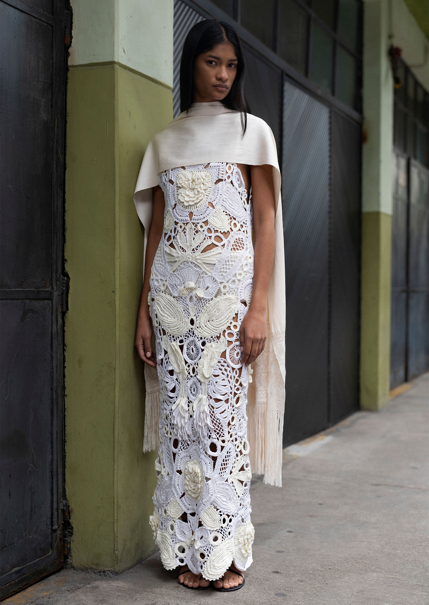 A woman wearing a white crochet dress with a white fringe silk shawl standing in a concrete hallway.