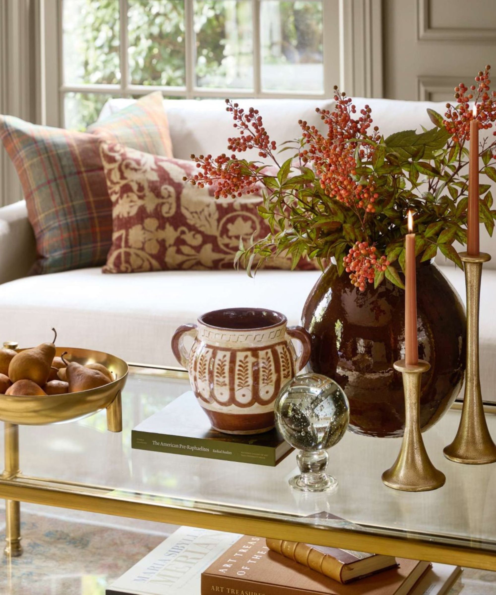 A gold and glass coffee table decorated for fall with gold candle sticks, ceramic vases and stacks of books