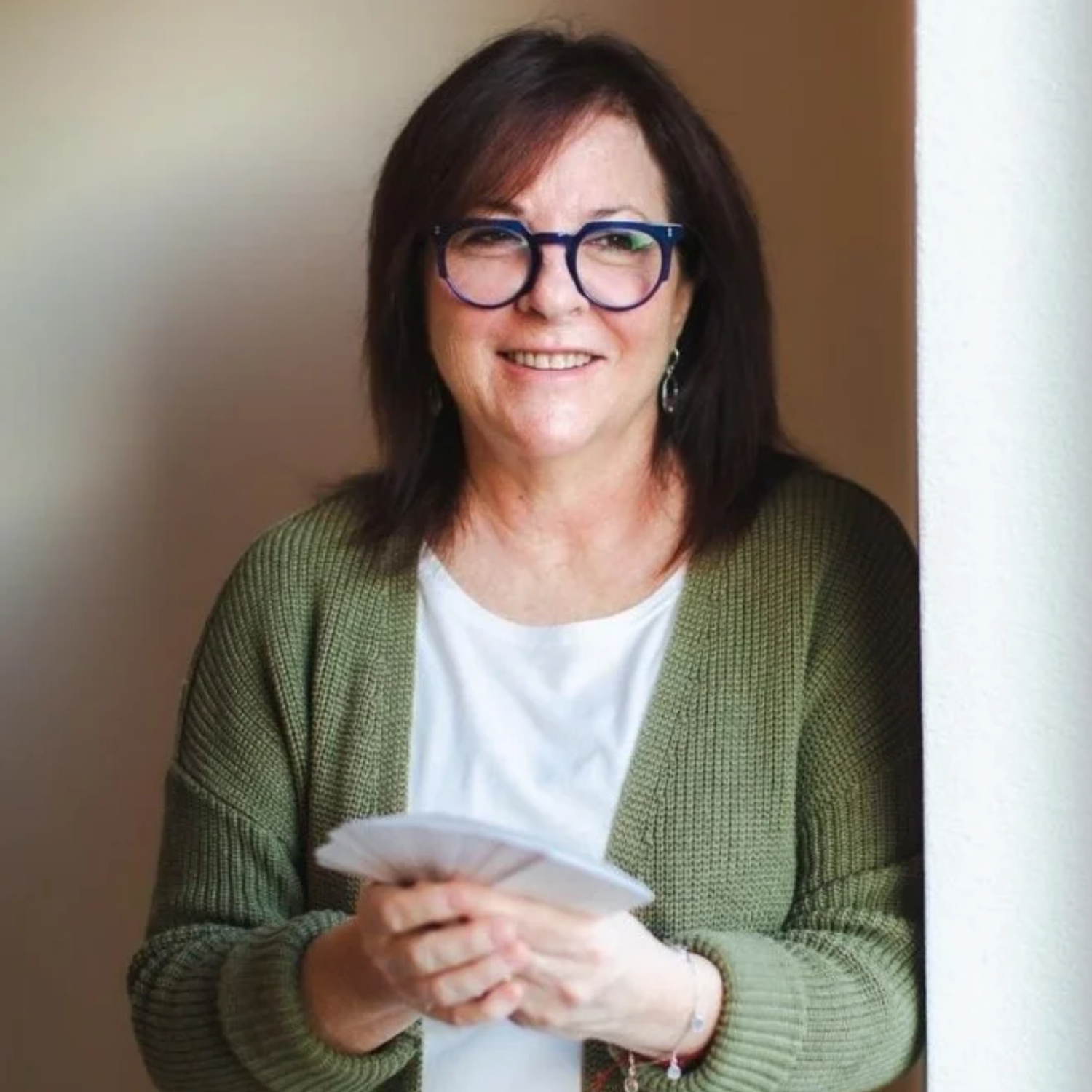 A headshot of a woman with short brown hair in a pair of blue glasses, dressed in a white round-neck t-shirt and a green woven cardigan