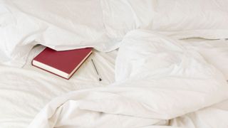A red diary tucked underneath a white pillow on a white bed with a pen lying next to it