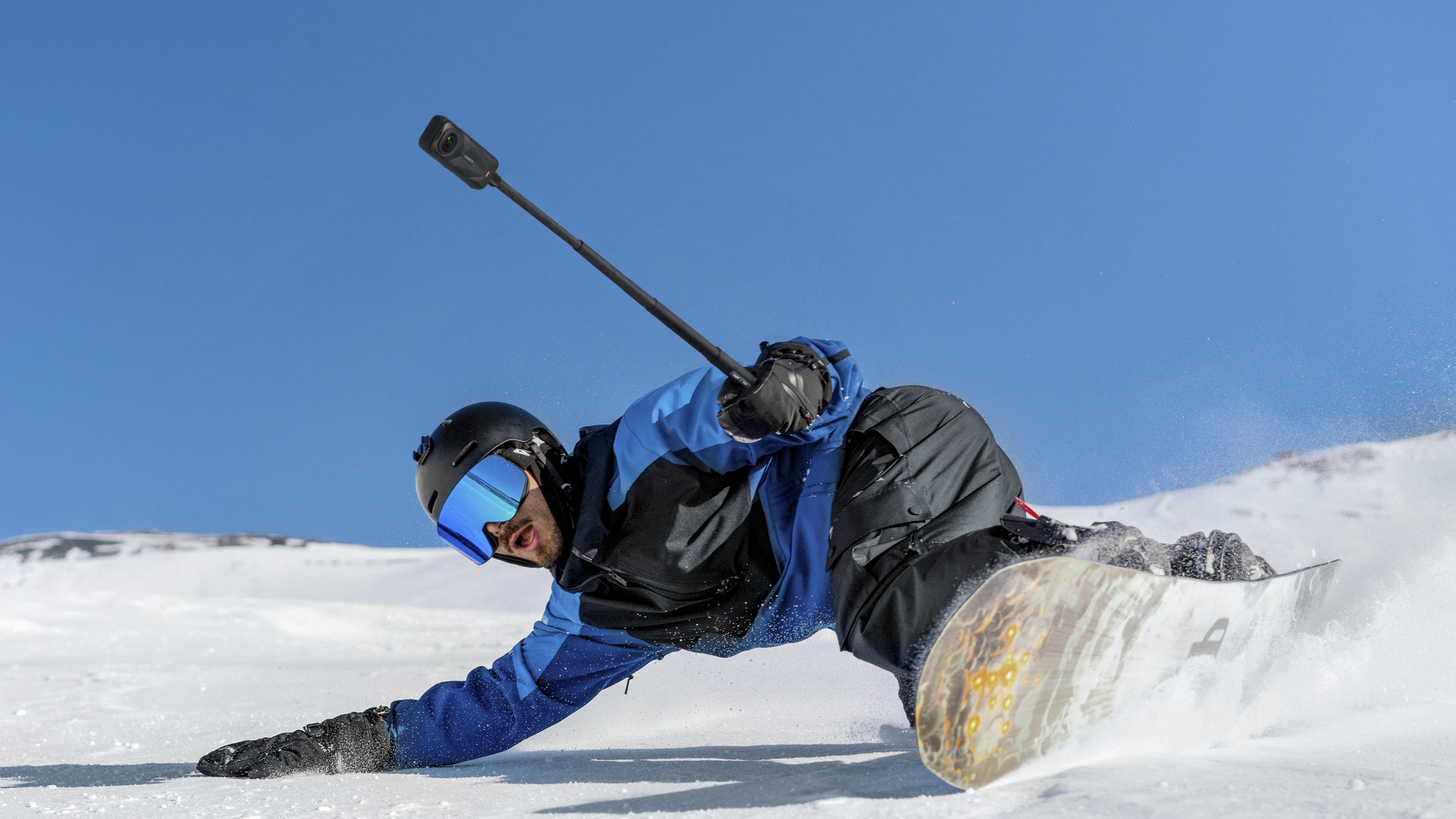 Insta360 X5 360 camera on a selfie stick, held by a snowboarder carving a turn, low to the powder