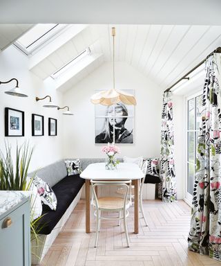Bright, white kitchen with paneled vaulted ceiling, wooden dining table with bench seating, floral curtains, wooden herringbone style flooring, wall lights, low hanging pendant over table