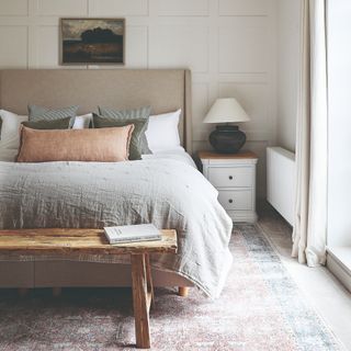 A neutral-coloured bedroom with wall panelling, natural bed linen and a faded Moroccan rug