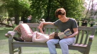 A peaceful moment from Notting Hill, where William sits on a park bench reading a book, while Anna rests her head in his lap, eyes closed, surrounded by lush greenery.