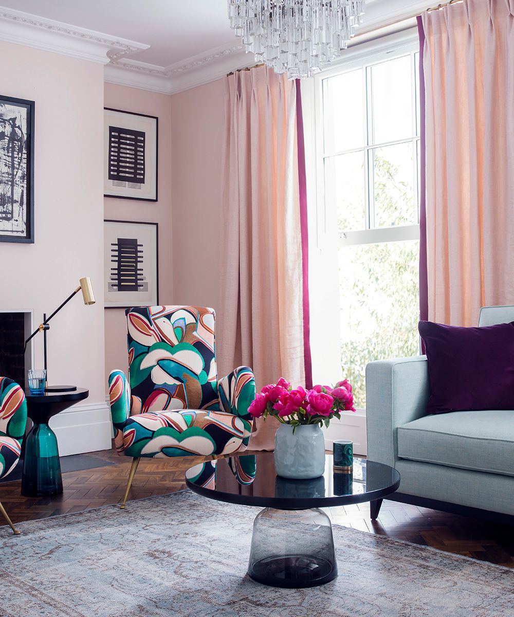 A pink living room with glass chandelier and abstract floral print chairs.