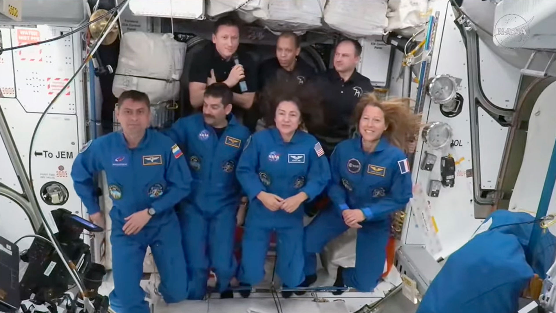 four people in blue flight suits and three in black polo shirts gather together aboard a space station