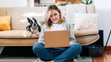 A smiling woman sits on the floor using a laptop with a cute dog beside her