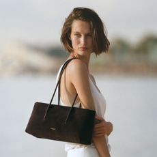 Woman wears white top, white trousers, and carries brown suede DeMellier Stockholm Shoulder bag