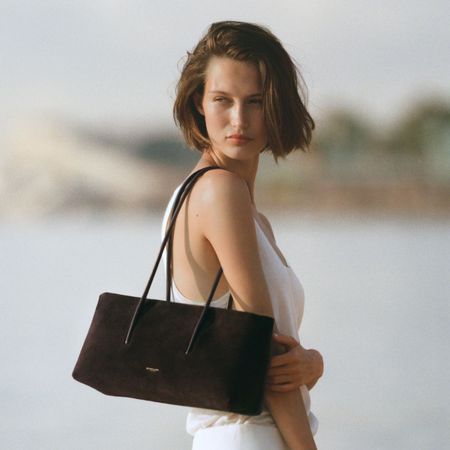 Woman wears white top, white trousers, and carries brown suede DeMellier Stockholm Shoulder bag