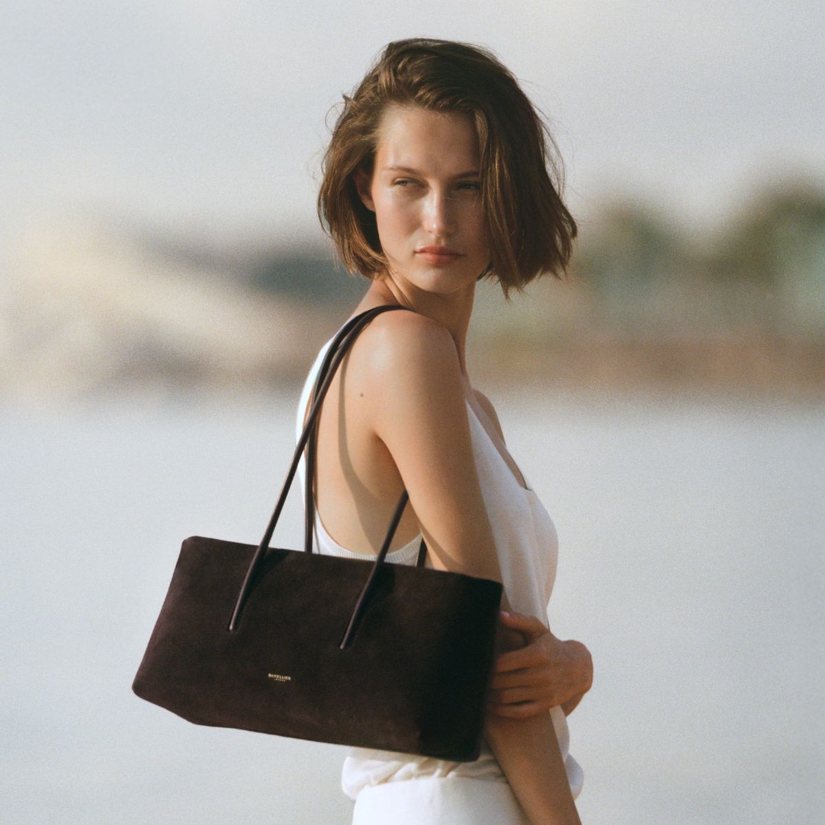 Woman wears white top, white trousers, and carries brown suede DeMellier Stockholm Shoulder bag