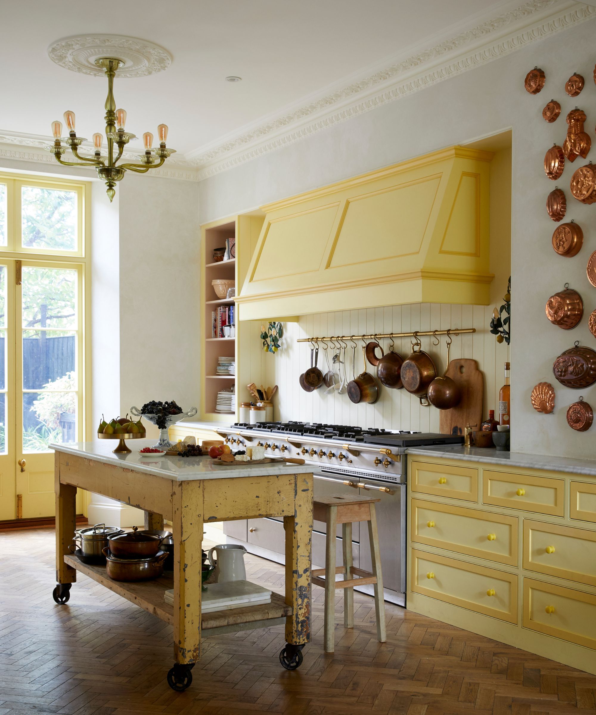 A yellow and white kitchen with brown parquet flooring. To the right are yellow cabinets, a silver hob, a white panelled wall with hanging copper pans, and a yellow range hood above. To the right are hanging copper pans. In front of the hob is a yellow island with a white countertop on wheels, underneath a green pendant lamp. Behind is a bright window.