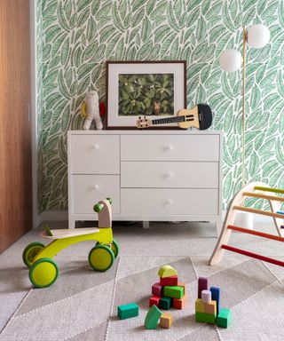 Green leaf print wallpaper in a gender neutral nursery idea with white furniture and colorful wooden toys.