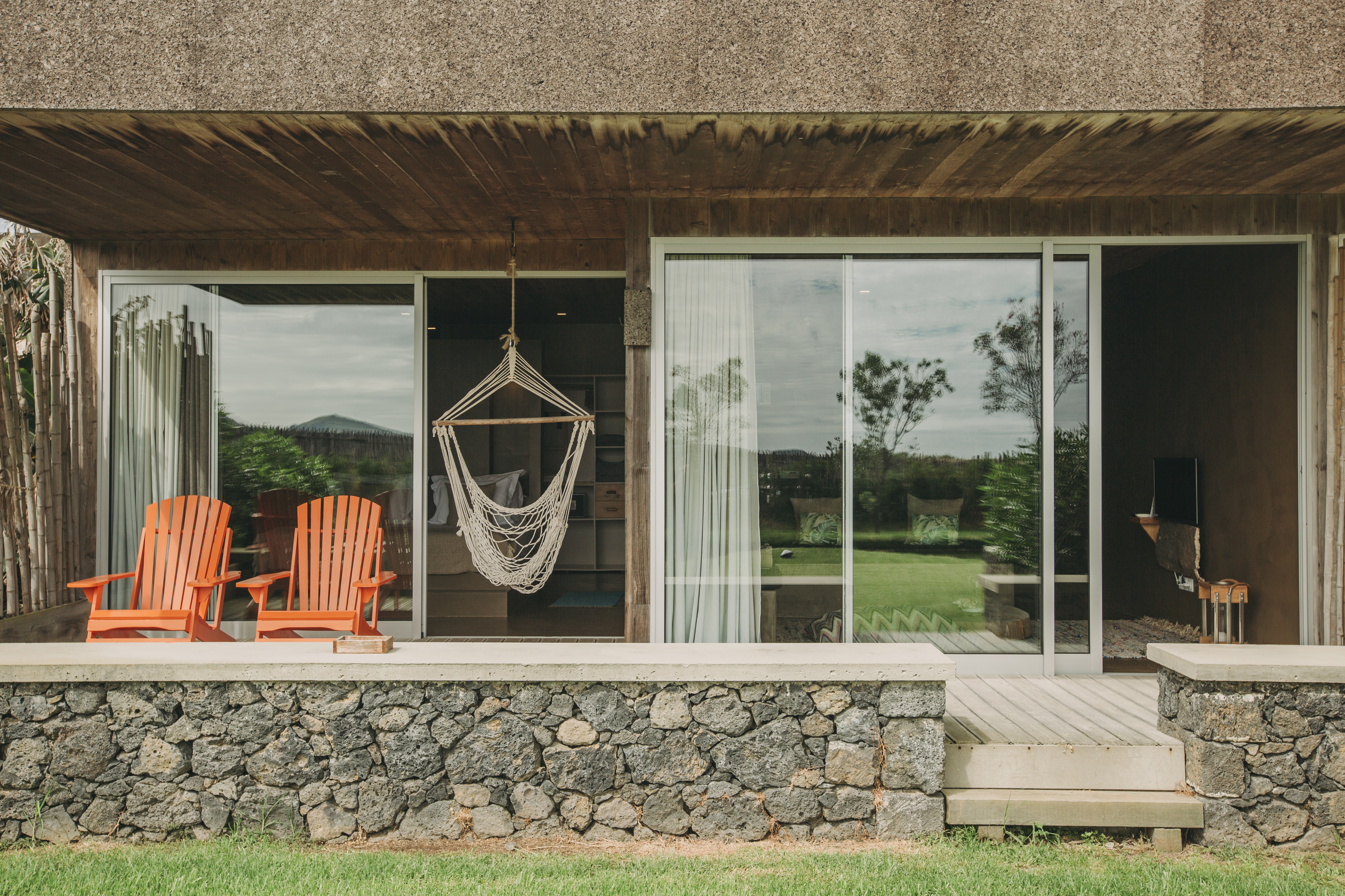 The brutalist exterior of an eco-retreat boasts cement, stone, and wooden details brought to life by colorful, terracotta deck chairs, whimsical furniture, and floor-to-ceiling windows.