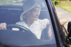 A woman in her eighties or nineties looks out the window from the driver's side of a car. She is wearing a hat and pearls.