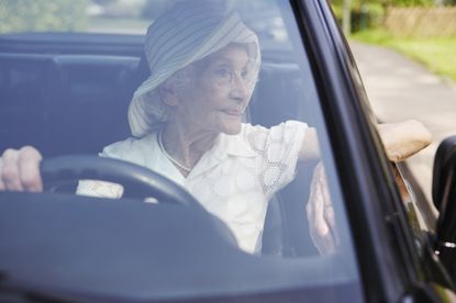 A woman in her eighties or nineties looks out the window from the driver's side of a car. She is wearing a hat and pearls.