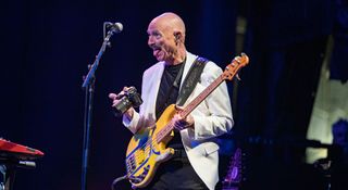 Tony Levin sticks his tongue out as he asks the audience to say cheese for a photo. He has a camera in one hand and his bass strapped around his neck.