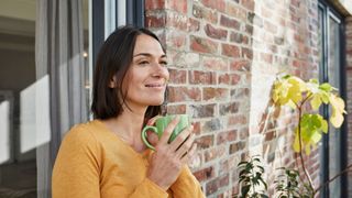 Woman holding green mug and smiling, standing in the doorway of a garden next to brick wall
