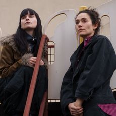 A woman in a fur-lined coat (Lola Petticrew as Catherine) sits on a kid's playground slide as another woman (Emmy Rossum as Alice) stands next to her, in Hulu's 'Furious.'