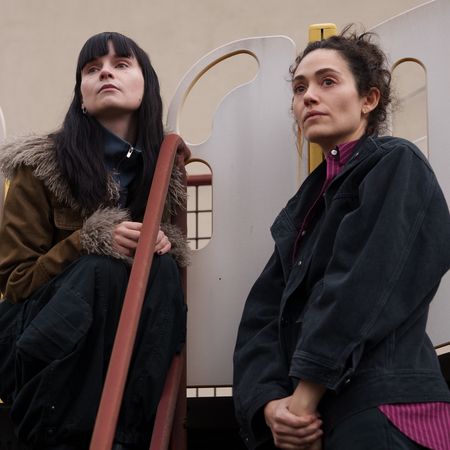 A woman in a fur-lined coat (Lola Petticrew as Catherine) sits on a kid's playground slide as another woman (Emmy Rossum as Alice) stands next to her, in Hulu's 'Furious.'